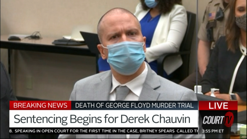 A white man in a surgical mask states into the camera
